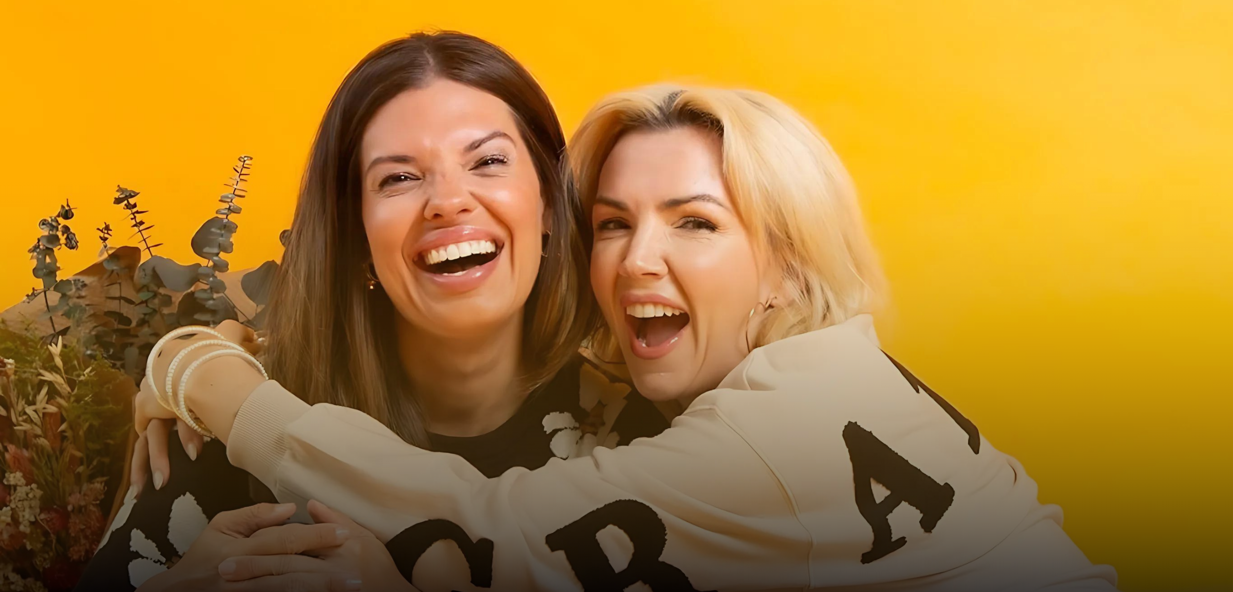 Two women laughing and hugging in front of a bright yellow background, both wearing cozy sweaters and radiating joy