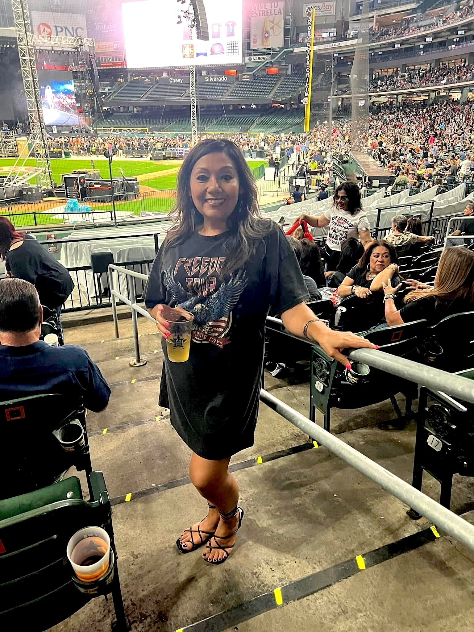 Woman smiling at a stadium concert, wearing a black oversized Freedom Tour t-shirt dress and strappy sandals, holding a drink and posing near the railing