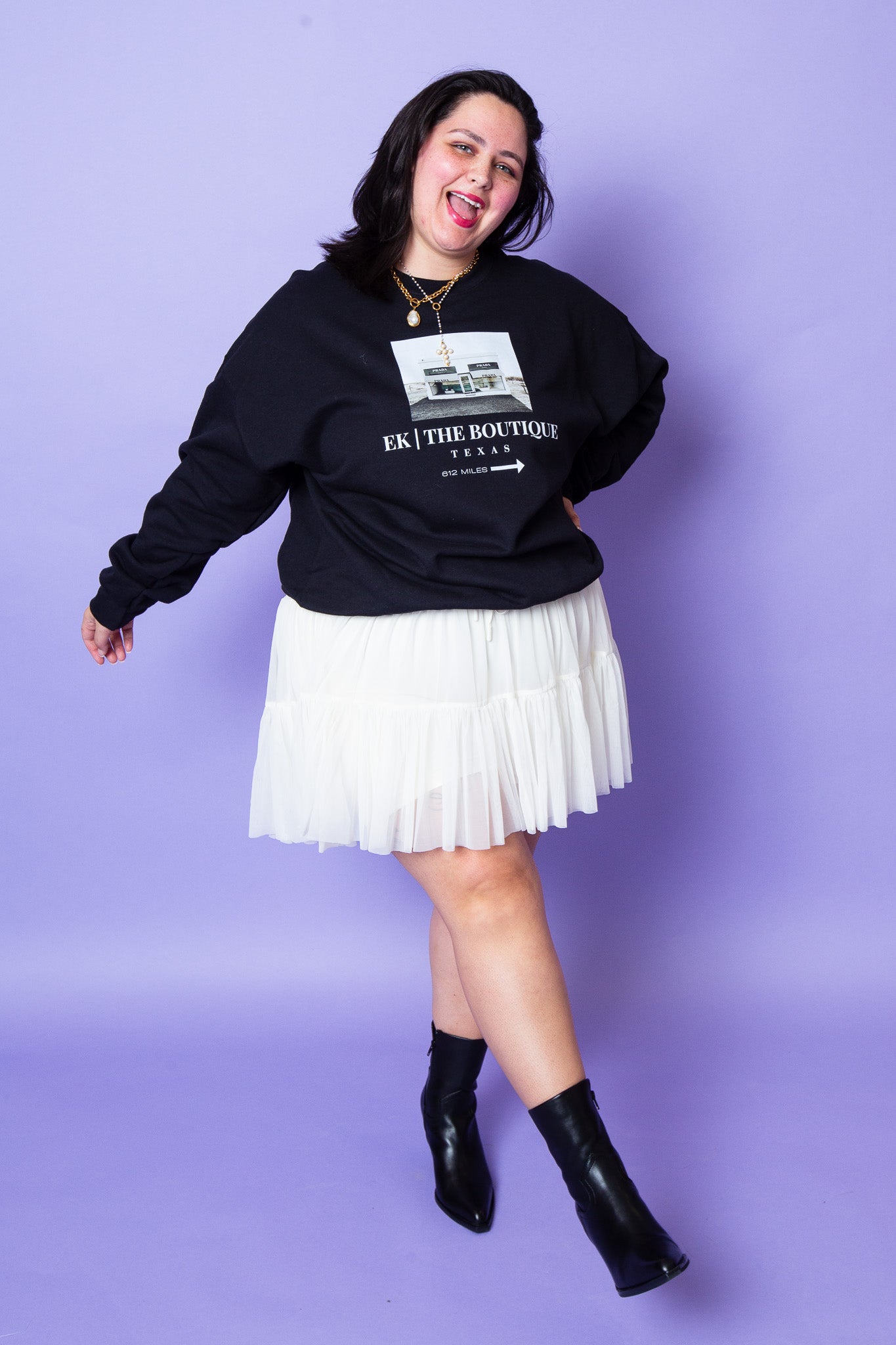 Confident woman smiling and posing playfully in a black EK | The Boutique Texas sweatshirt, white skirt, and black ankle boots against a lavender background