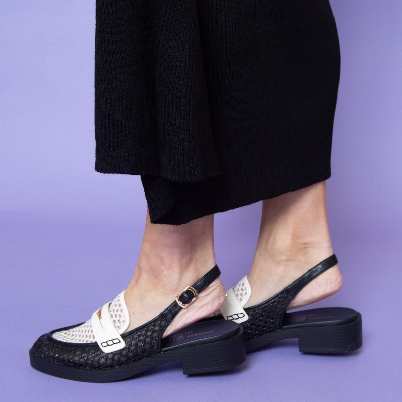Woman wearing black and white woven slingback loafers with block heels, styled with a black ribbed skirt on a lavender background