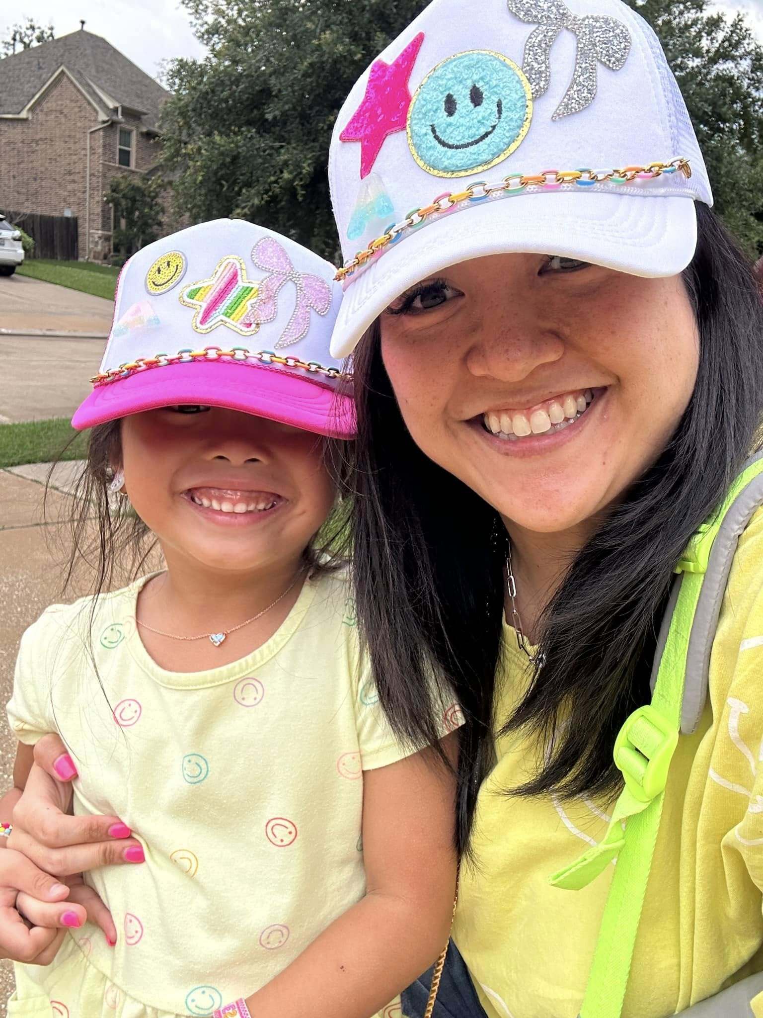 Smiling woman and young girl wearing matching decorated trucker hats and yellow outfits with smiley face designs, posing together outdoors
