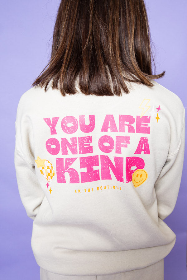 A person with shoulder-length brown hair is seen from the back wearing a cream-colored sweatshirt. The sweatshirt features a bold, pink graphic design with the words "YOU ARE ONE OF A KIND" along with small decorative elements like a yellow smiley face, a lightning bolt, and sparkles. Below the main text, "EK THE BOUTIQUE" is printed in yellow. The background is a soft purple color.