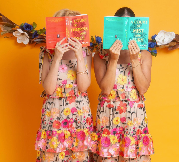 Two women wearing matching floral dresses standing in front of an orange background, holding up books from the A Court of Thorns and Roses series to cover their faces