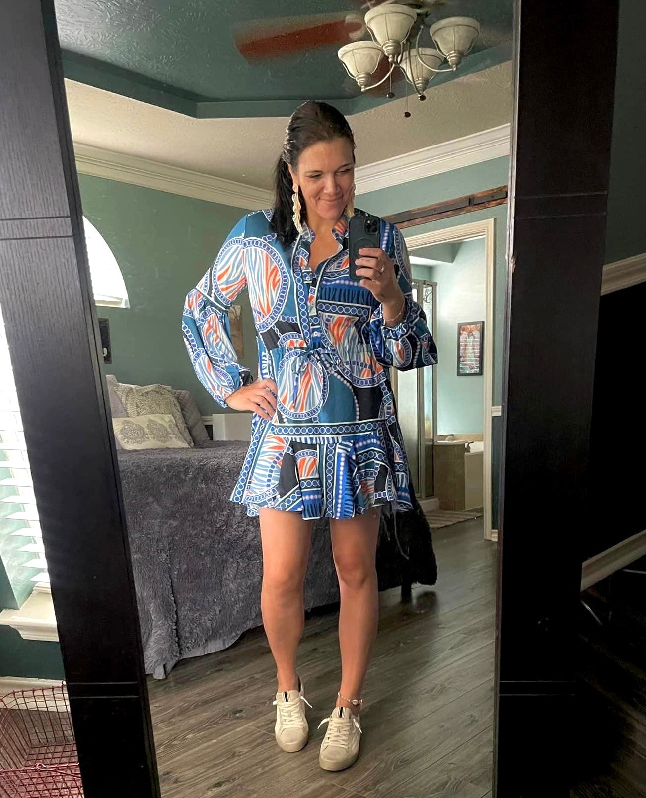 Woman taking a mirror selfie in a colorful patterned dress with long sleeves and white sneakers, smiling confidently in a bedroom