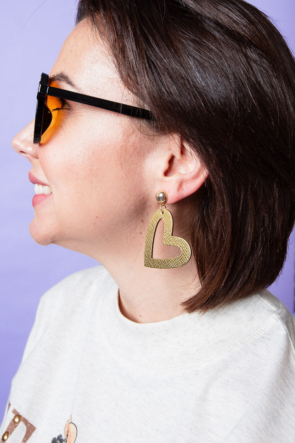 Smiling woman wearing oversized yellow-tinted sunglasses and a gold textured open-heart statement earring against a lavender background