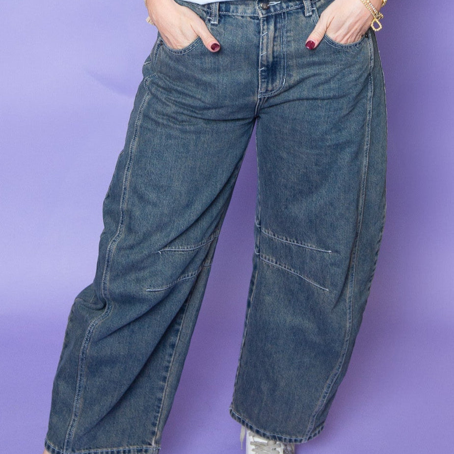 Close-up of a person wearing high-waisted, wide-leg denim jeans with unique angled seam details, standing against a lavender background